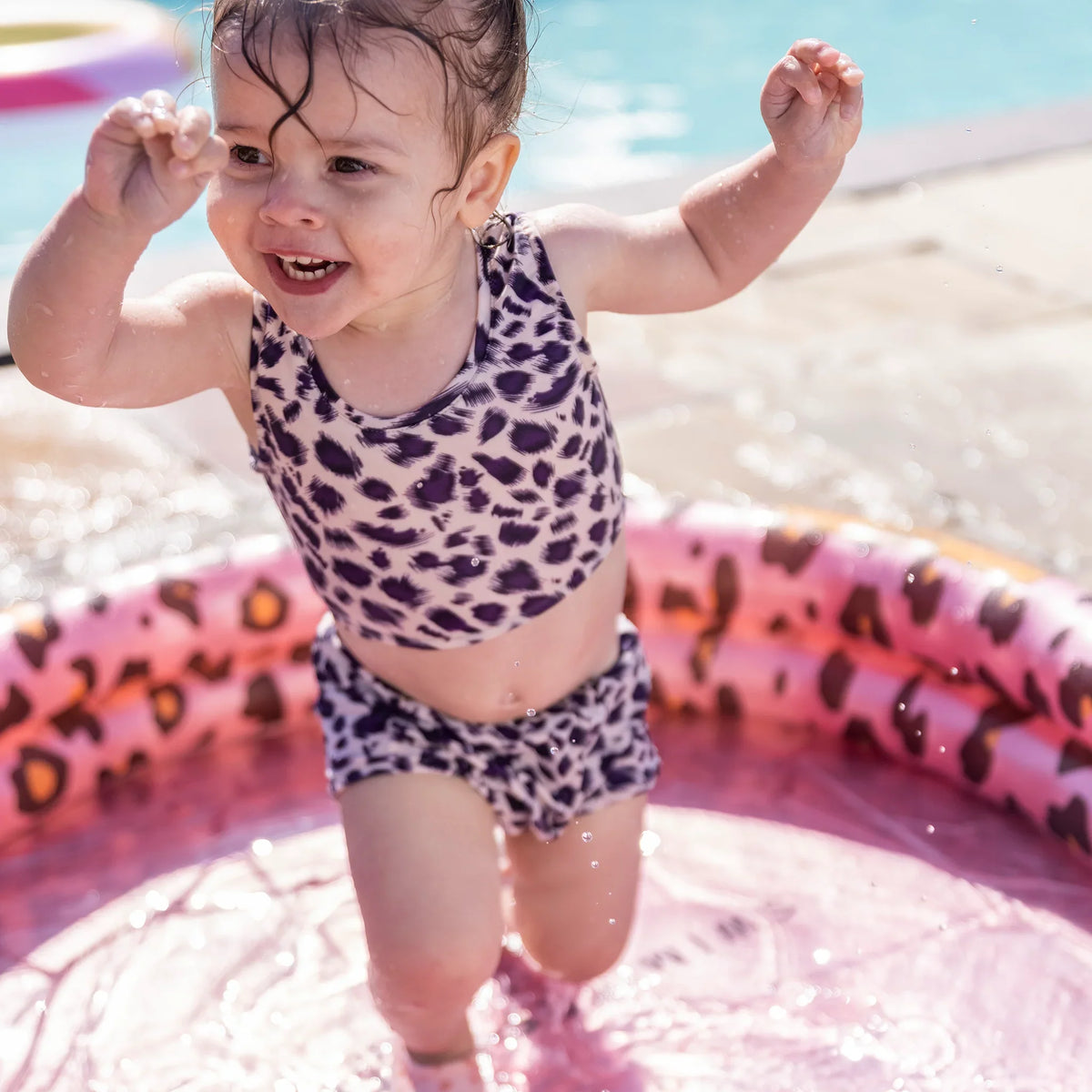 Kind staat met opgeheven armen in een roze babyzwembad met luipaardprint.