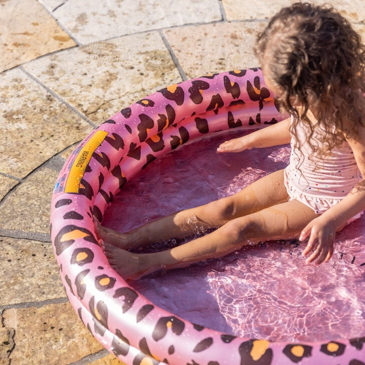 Kind zit met gestrekte benen in een roze babyzwembad met luipaardprint op een stenen terras.