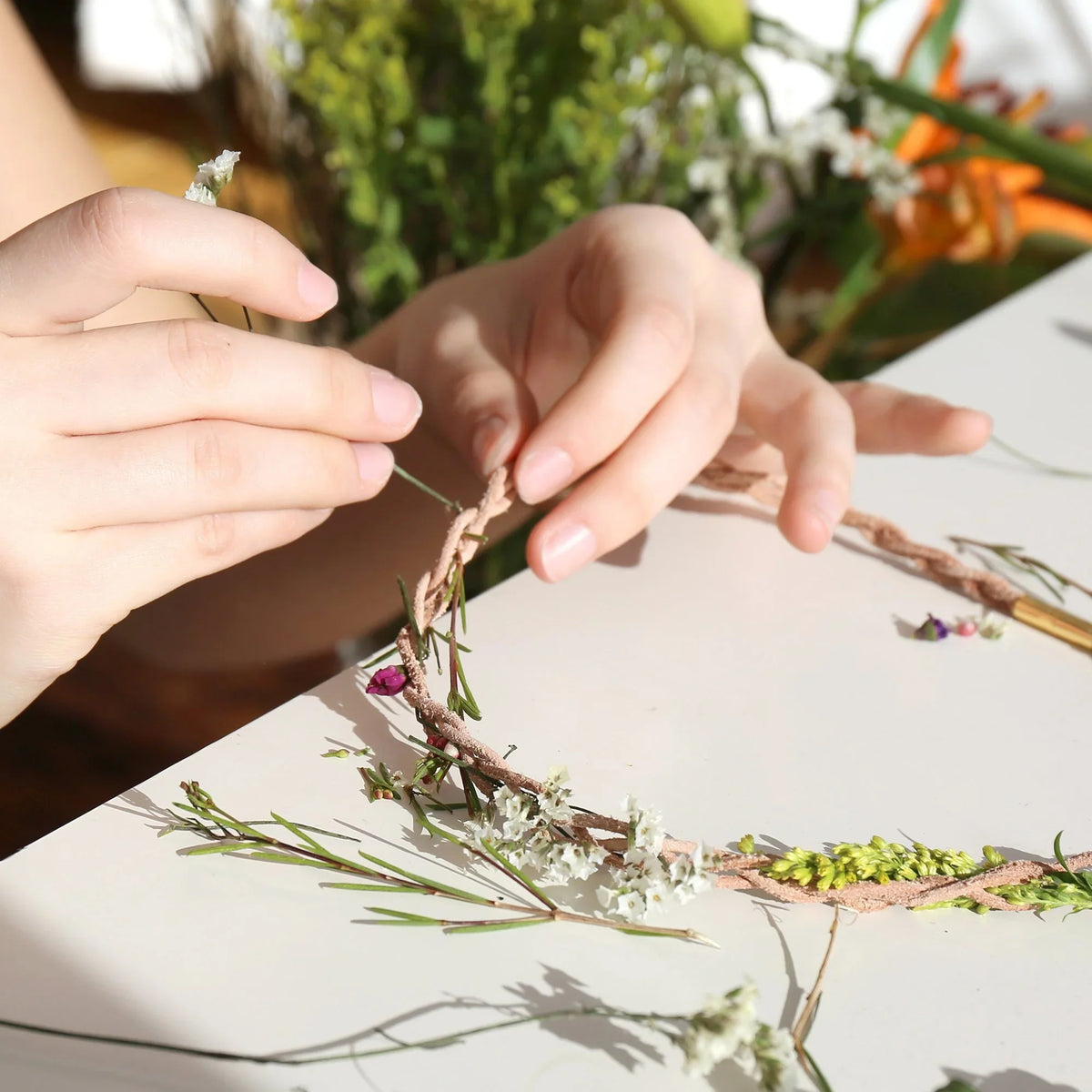 Handen bevestigen bloemen en groene takjes aan een bloemenketting op een witte tafel.