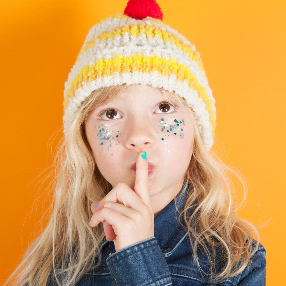 Kind met turquoise nagellak houdt een vinger voor de lippen, met glitters op de wangen en een gebreide muts.