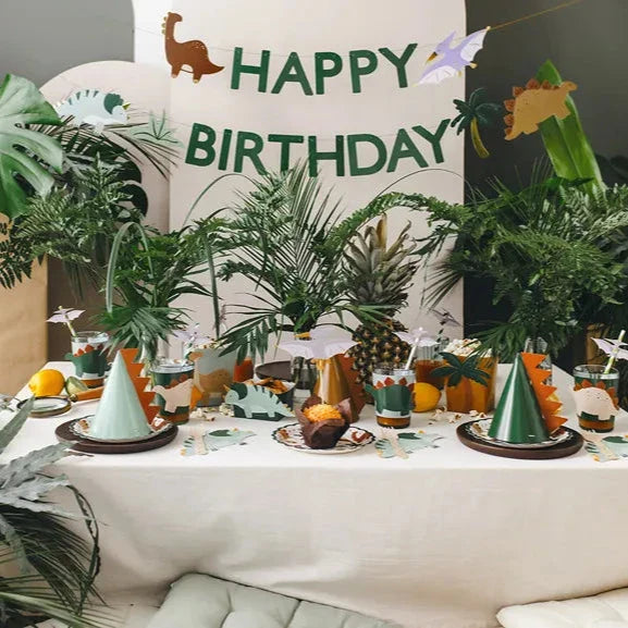 Dino-feesthoedjes op een gedekte verjaardagstafel met dinosaurusdecoraties, planten en een verjaardagsslinger.