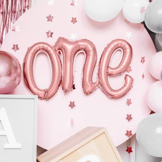 Roségouden folieballon met het woord 'one' tegen een roze achtergrond met sterren en ballonnen.