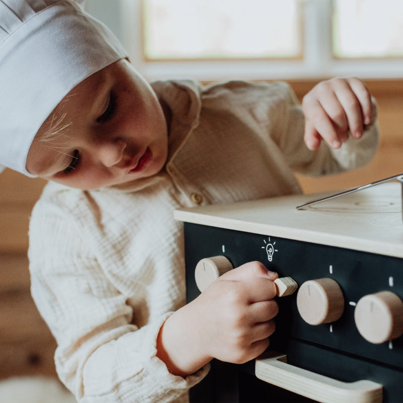 Kind met koksmuts bedient de houten schakelaar op de zwarte houten speelkeuken.