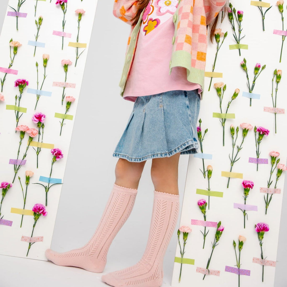 Kind met lichtroze Lauren Pink kniekousen, denimrok en kleurrijk vest voor een wand met bloemen.