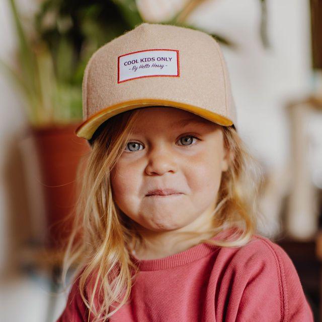 Vooraanzicht van een kind met de beige Melton Creamy pet met okerkleurige klep en rood omrand embleem.