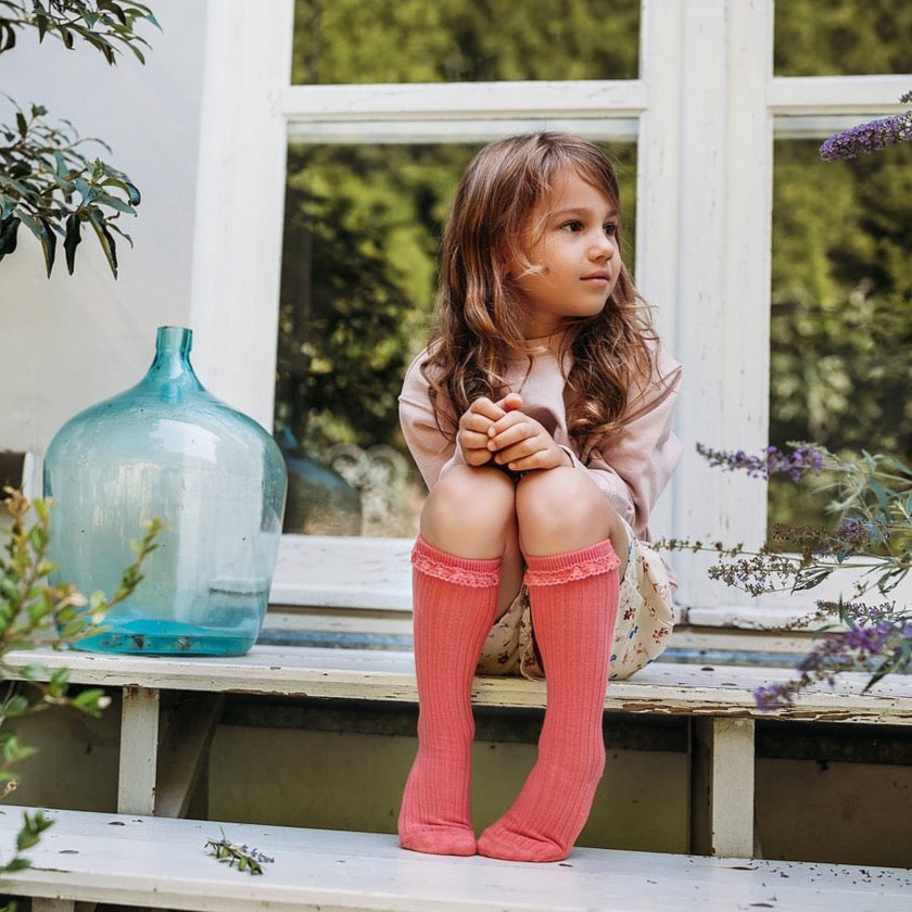 Kind met koraalkleurige Mono Baby kniekousen met kanten randjes, zittend voor een raam naast een blauwe vaas.