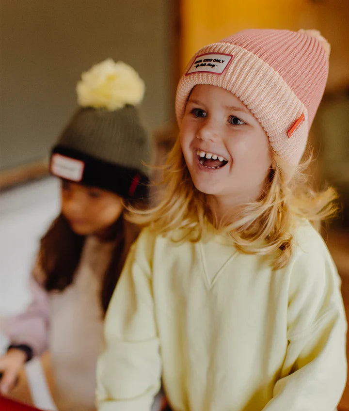 Lachend kind draagt de roze Hello Hossy Block Blush muts met lichtroze omslag en label vooraan.