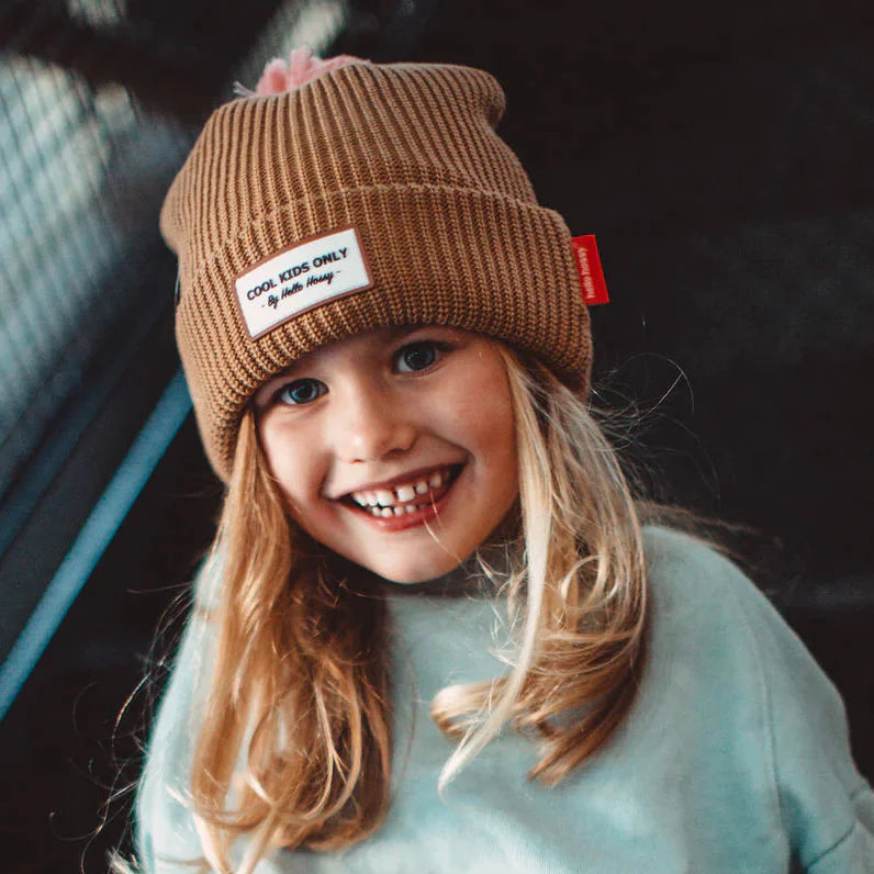 Lachend kind met bruine Block Macaroon muts van Hello Hossy, met omgeslagen rand en roze pompon.