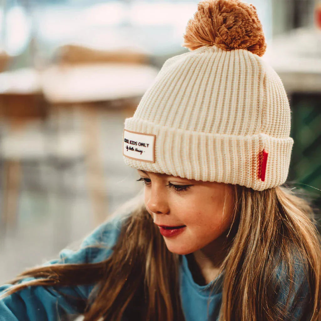 Kind met de crèmekleurige Muts Block Sandy van Hello Hossy met bruine pompon en omgeslagen rand.