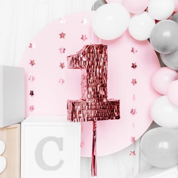 Roségouden piñata in de vorm van het cijfer 1 voor een roze achtergrond met sterren en ballonnen.