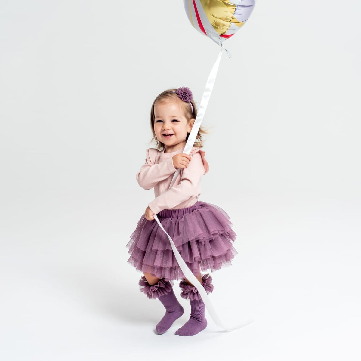 Kind met de oudspaarse Tutu Bloomer, een lichtroze top en paarse kniekousen, met een ballonlint in de hand.