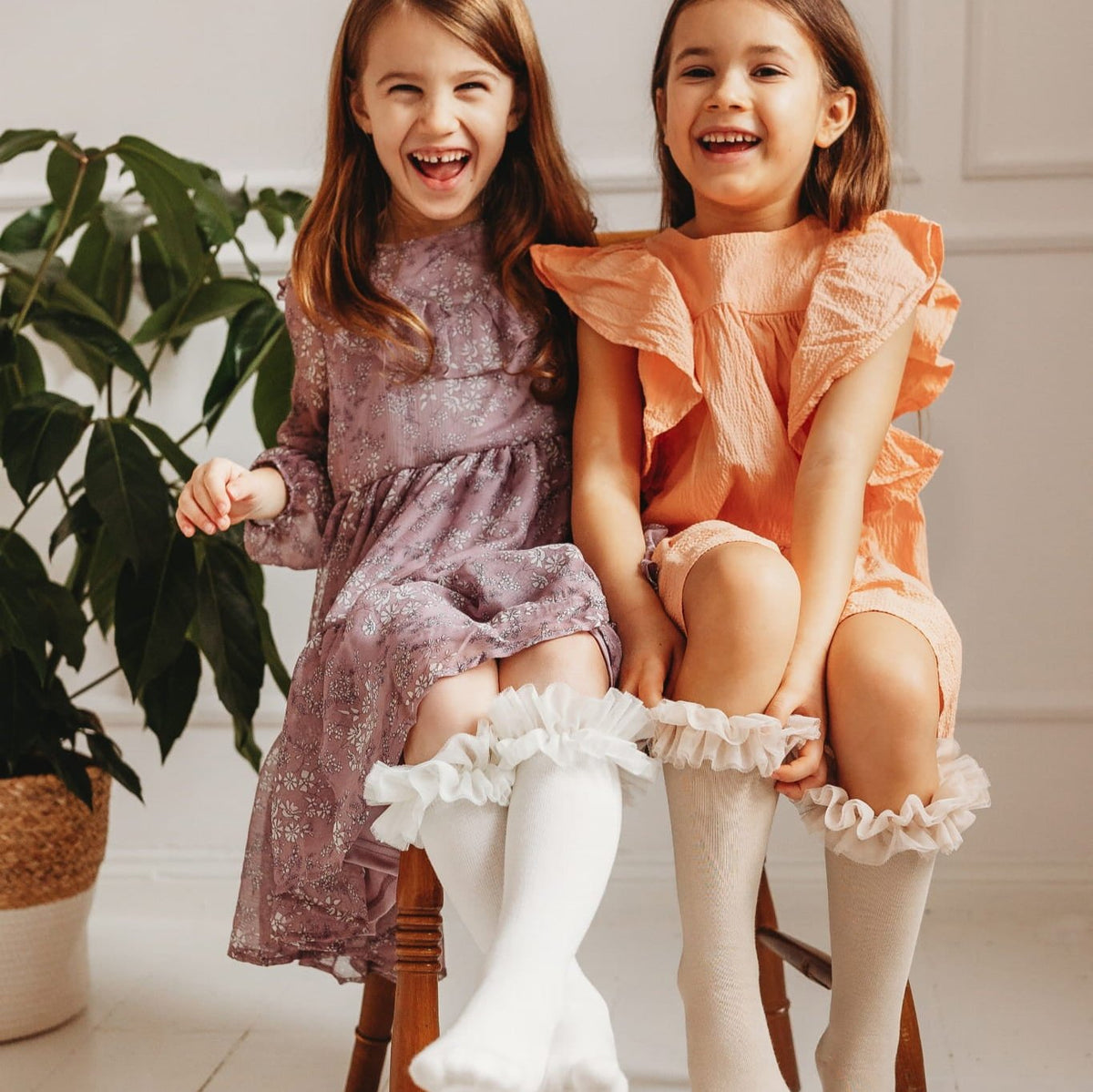 Twee lachende kinderen op een houten stoel, met witte en beige tutu kniekousen onder hun jurken.