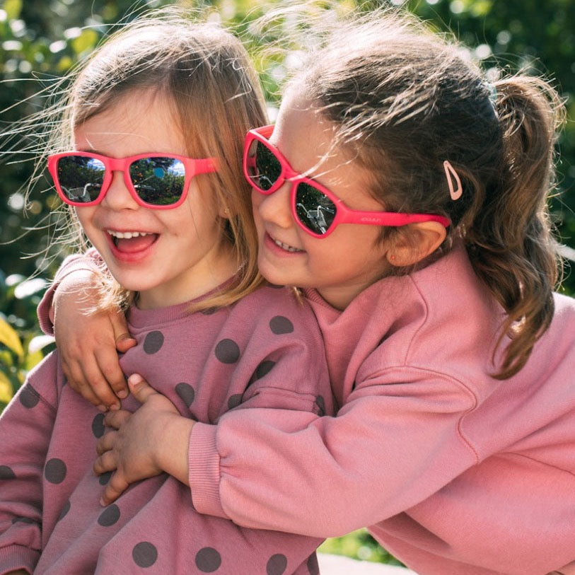 Twee kinderen met een roze Aspen zonnebril Camellia Rose omhelzen elkaar buiten.