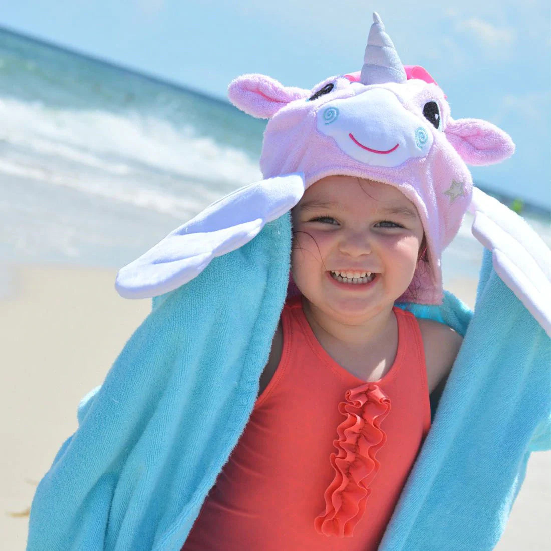 Kind op het strand met turquoise Zoocchini badcape en roze eenhoorncapuchon, met de armen omhoog.