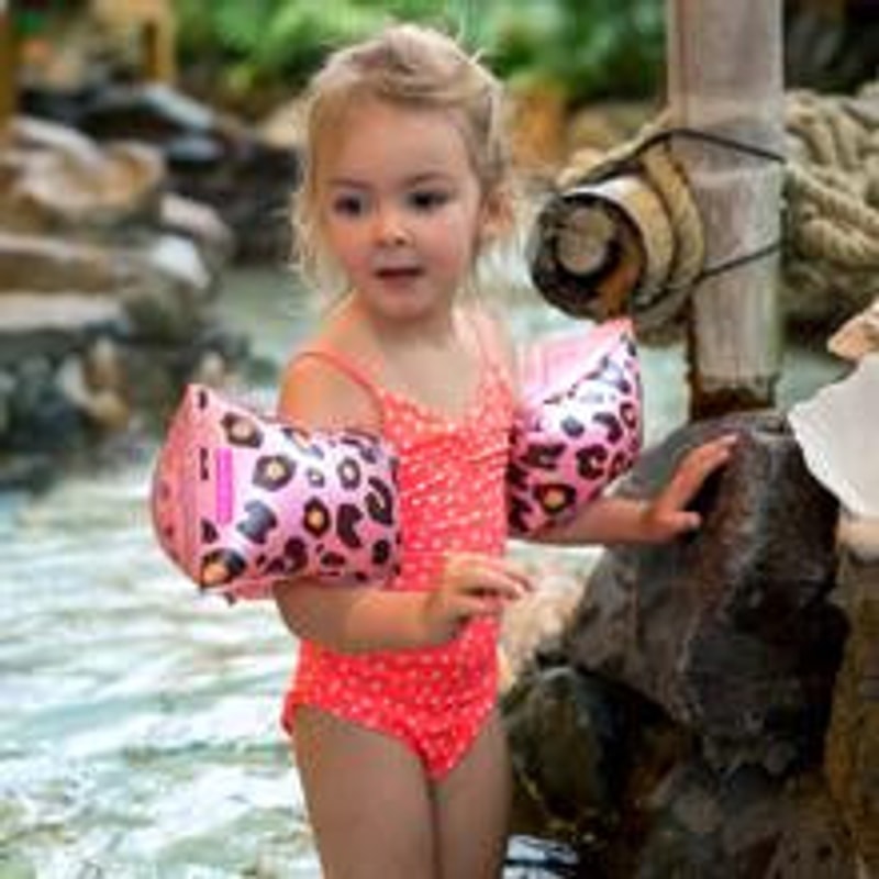 Meisje in een koraalroze badpak met roze panterprint zwembandjes staat in het water bij rotsen.