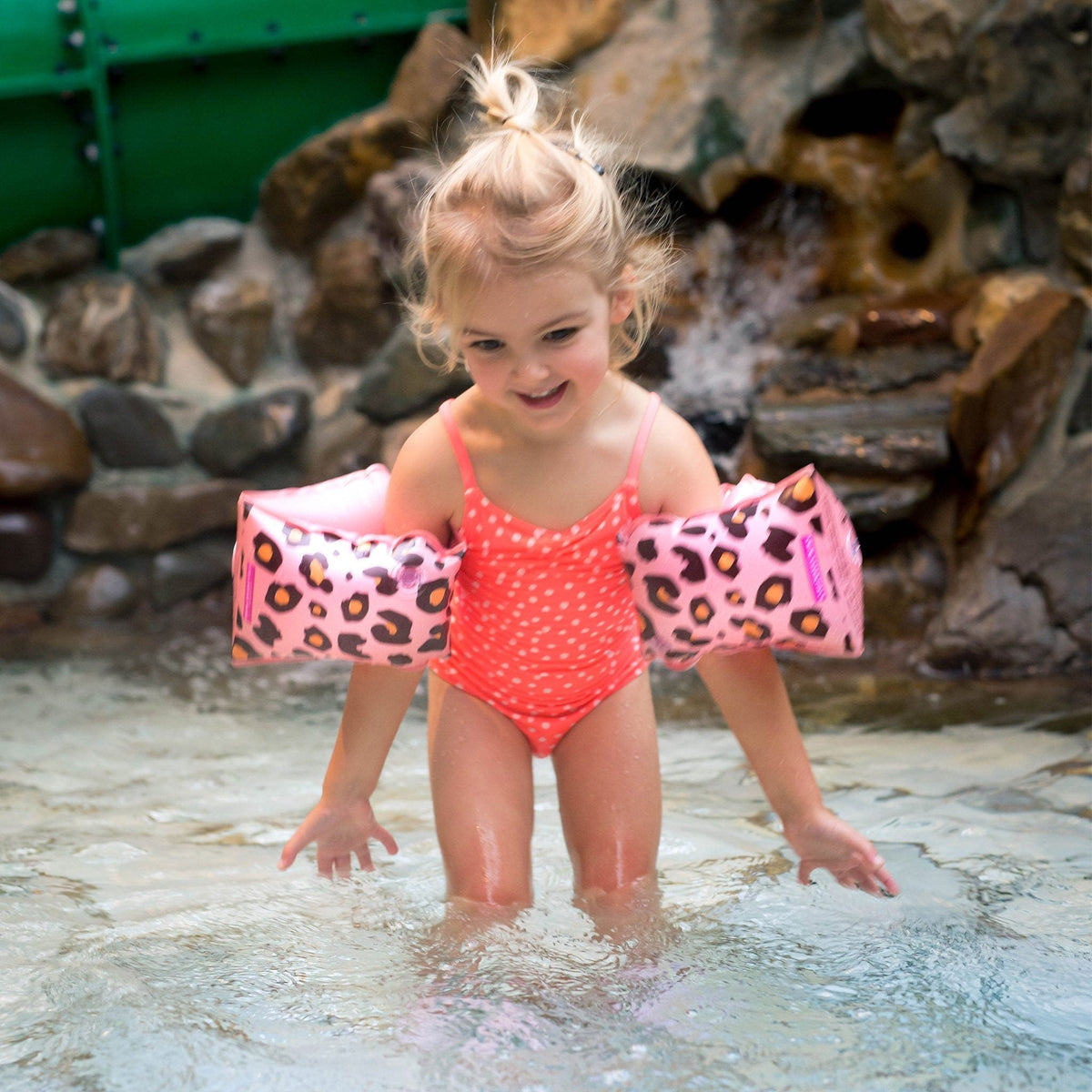 Meisje met roze panterprint zwembandjes en een koraalroze badpak speelt in ondiep water.