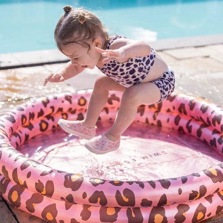 Kind springt in een roze babyzwembad met luipaardprint naast een groot zwembad.