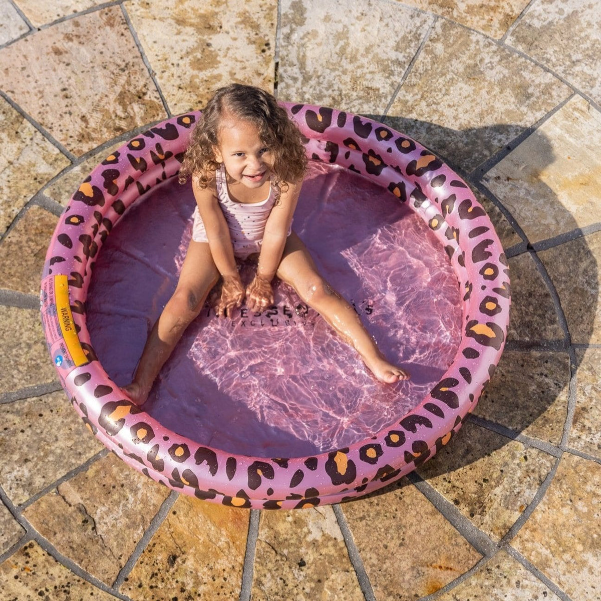Bovenaanzicht van een roze babyzwembad met luipaardprint en een zittend kind op een stenen terras.