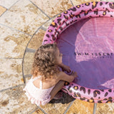 Kind hurkt naast een roze babyzwembad met luipaardprint en houdt de opblaasrand vast.