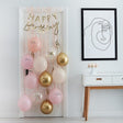 Ballonnen deurkit met roze, perzikkleurige en gouden ballonnen en gouden Happy Birthday-slinger aan een witte deur.