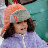 Close-up van een kind met de oranje en grijsgroene Cozy Jim wintermuts met oorkleppen in de sneeuw.