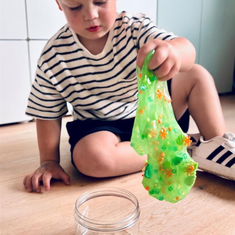 Een kind zit op de vloer en rekt groen Dino Dew-slijm met bedeltjes uit boven een open pot.