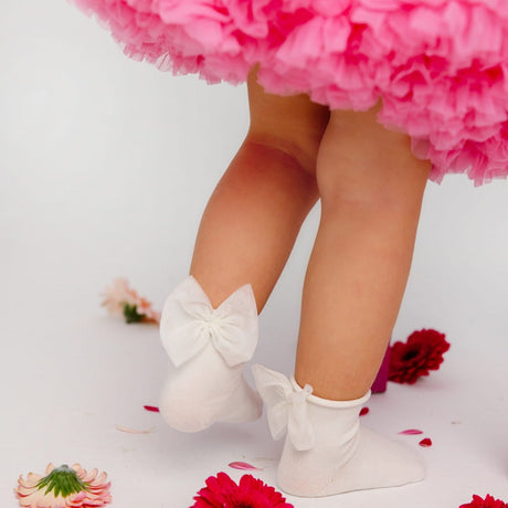 Kind draagt roomwitte Daisy sokken met grote strikken onder een roze rok, met bloemen op de grond.