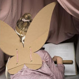 Kind van achteren met bruine kartonnen feeënvleugels en een tiara, zittend voor een open boek.