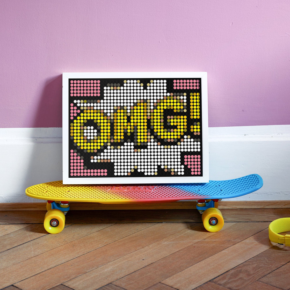 Dot On omg stickerposter in een witte lijst op een kleurrijk skateboard tegen een roze muur