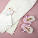 Roze eenhoornpantoffels naast een witte deken en een wit eenhoornknuffeldoekje op een roze ondergrond.