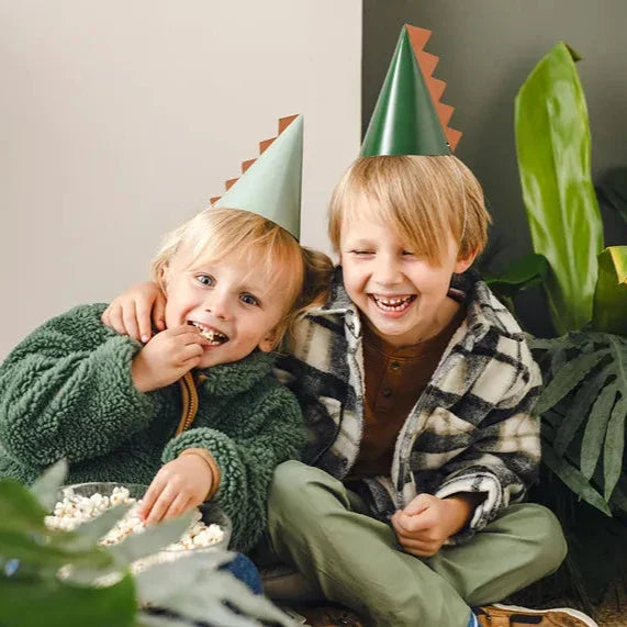 Twee lachende kinderen dragen lichtgroene en donkergroene dino-feesthoedjes met oranje stekels.
