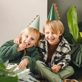 Twee lachende kinderen dragen lichtgroene en donkergroene dino-feesthoedjes met oranje stekels.