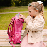 Zijaanzicht van de roze Flashbag Lou-rugzak op een houten bank terwijl een kind de bovenklep vasthoudt.