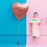 Roségouden hartvormige folieballon aan een lint naast een vrouw, tegen een turquoise en roze muur
