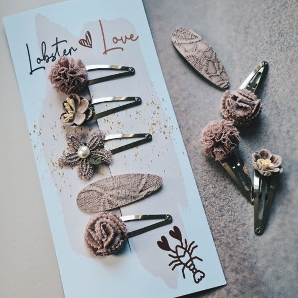 Haarspeldjes Misty Mauve met bloemen en ovale vormen in mauve tinten, op een kaartje en een grijze ondergrond.