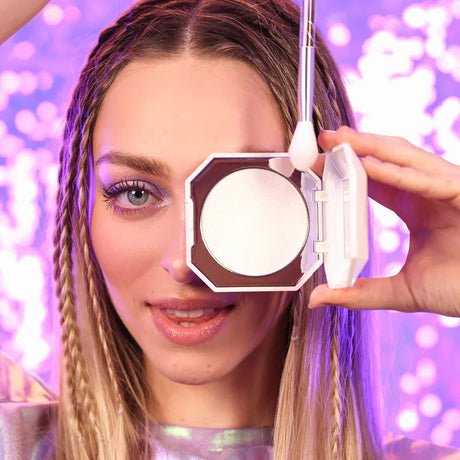 Vrouw houdt een open highlighterdoosje en highlighterkwast Camille voor haar gezicht tegen een paarse achtergrond.