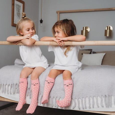 Twee kinderen op een bed met roze Lilly a Pinky Kitty kniekousen met witte hartjes en kattengezichtjes.