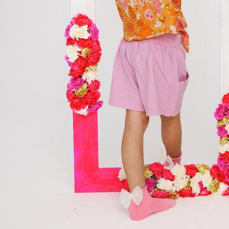 Kind draagt roze Magnolia sokken met witte strikken, een roze short en een gebloemde top bij een bloemenspiegel.
