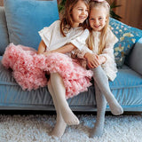 Twee meisjes op een blauwe bank dragen de maillot Josephine in beige en grijs onder een roze rok en witte jurk.