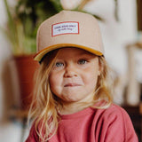 Vooraanzicht van een kind met de beige Melton Creamy pet met okerkleurige klep en rood omrand embleem.