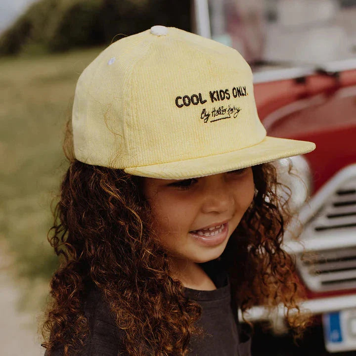 Kind met de gele Mini Citrus-pet van Hello Hossy met platte klep, bij een rode auto.