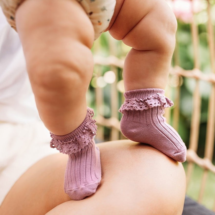 Baby draagt oudroze Mono Baby sokken Heather met ribstructuur en kanten ruches, staand op een bovenbeen.