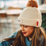 Kind met de crèmekleurige Muts Block Sandy van Hello Hossy met bruine pompon en omgeslagen rand.