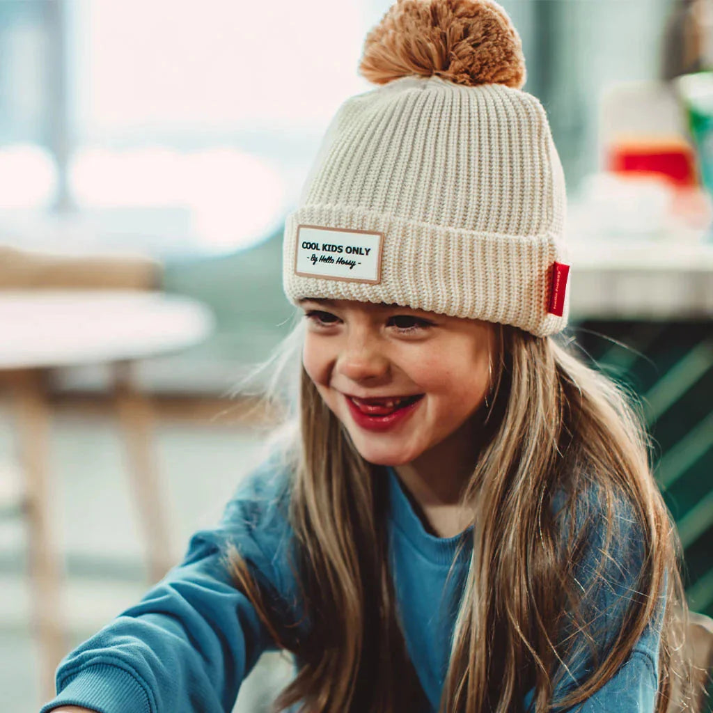Lachend kind met de crèmekleurige Muts Block Sandy van Hello Hossy met bruine pompon en label op de voorkant.