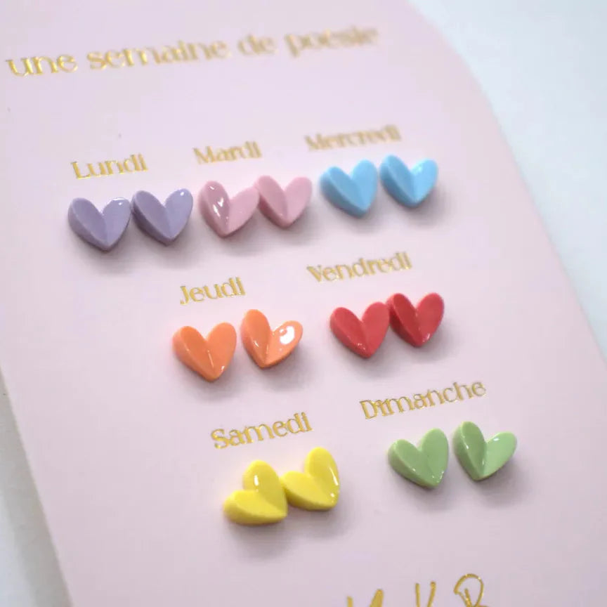 Detail van zeven paar kleurrijke hartjesoorbellen op een roze kaart met Franse weekdagen.