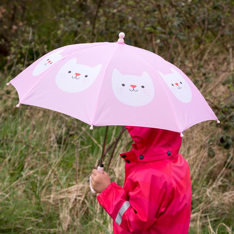 Kind in een rode regenjas met een open roze Cookie The Cat paraplu met witte kattenkoppen, buiten in het groen.