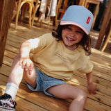 Kind met lichtblauwe pet Rubber Hortensia met roze klep, zittend op een houten vloer.