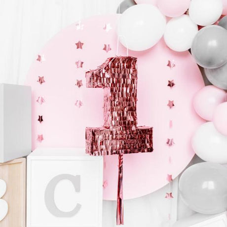 Roségouden piñata in de vorm van het cijfer 1 voor een roze achtergrond met sterren en ballonnen.