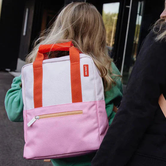 Kind draagt de lichtroze en roze Hello Hossy Mini Primrose rugzak met oranje handgrepen en voorvak met rits.