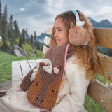 Meisje op een houten bank met de bruine Peggy Pony-sjaal met ponygezicht en roze bloem, en bijpassende oorwarmers.